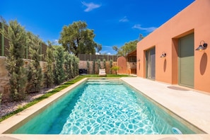 A rectangular swimming pool with clear water, surrounded by a stone wall and greenery, with a modern building on one side. - Villa Analidia Comfort (Koridallos)