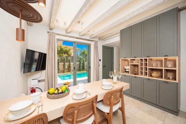 A modern dining area with a pool view, a flat-screen TV, and a kitchen with grey cabinets. - Villa Analidia Comfort (Koridallos)