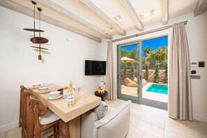 Un coin repas avec une table en bois, des chaises, une bouteille de vin et un verre. Un téléviseur est fixé au mur, et une porte-fenêtre coulissante donne accès à une zone avec piscine.