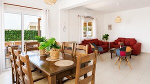 A dining area with a wooden table, chairs, and woven placemats. There is a red sofa and a small table with a vase of flowers. - Argiro Bright Villa - Four Bedroom Villa, Sleeps 8 (Pernera)