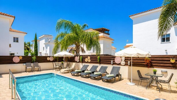 A swimming pool with lounge chairs and umbrellas, surrounded by white buildings and palm trees. - Argiro Bright Villa - Four Bedroom Villa, Sleeps 8 (Pernera)