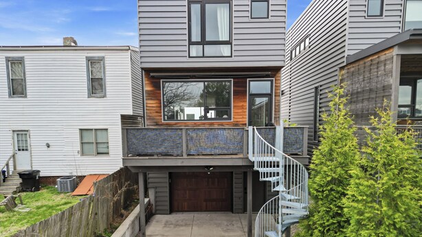 Ein modernes, zweistöckiges Haus mit einer überdachten Terrasse, einer Holzterrasse und einer Wendeltreppe, die zu einem Balkon führt.