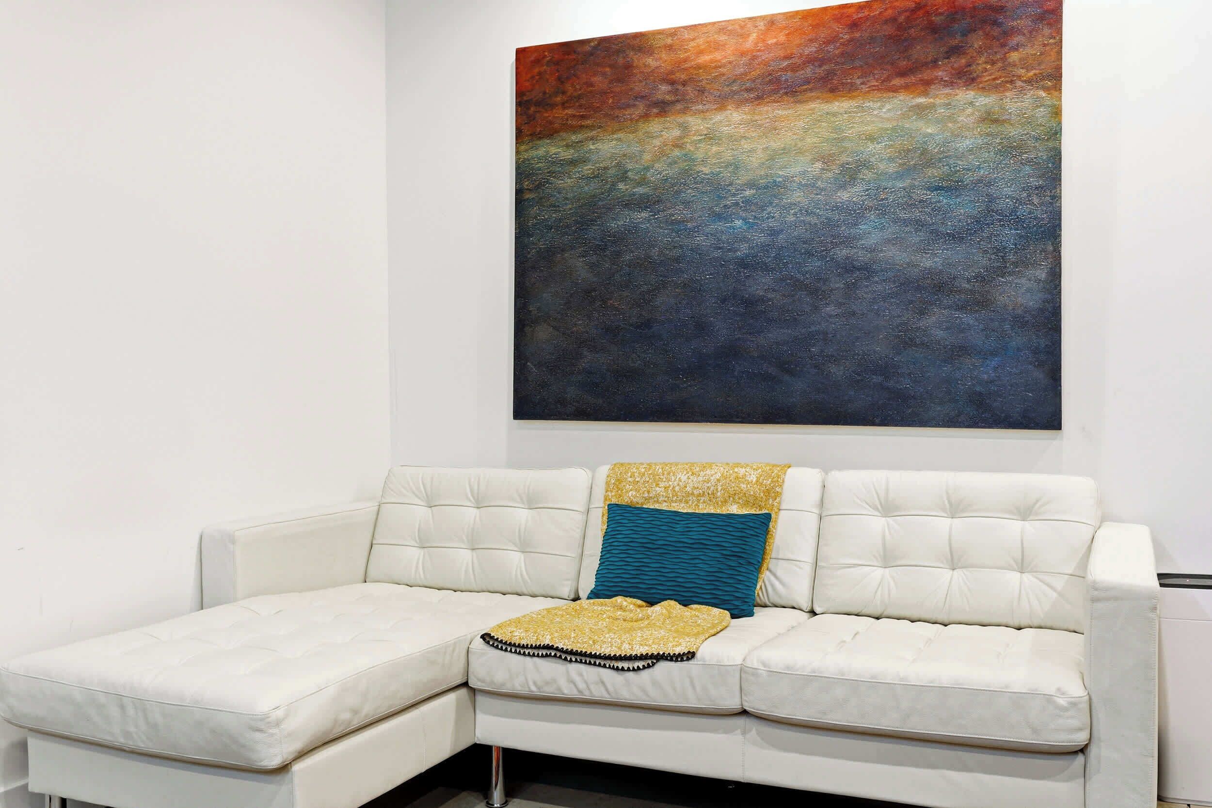 A white leather sofa with a gold and blue throw blanket, positioned in front of a large abstract painting.