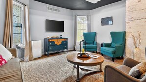 Ein Wohnzimmer mit einer Couch, einem Couchtisch, einem blaugrünen Sessel, einer blauen Kommode, einem Fernseher und einem Fenster mit Vorhängen.