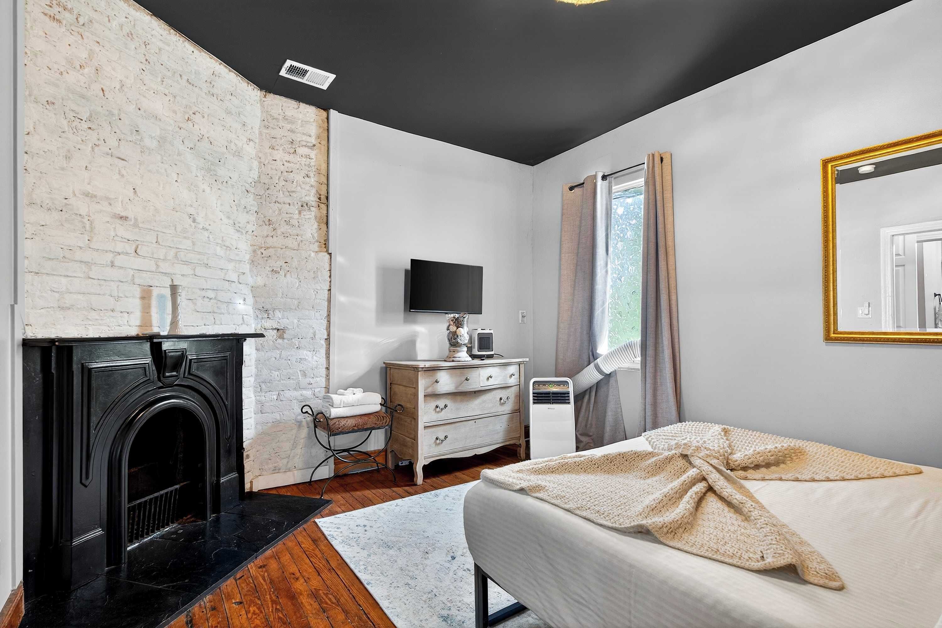 A bedroom with a fireplace, a bed, a dresser, a television, and a window with curtains.