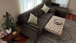 A living room with a sectional sofa, ottoman, and a patterned rug. - Charming Stylish & Cozy Coastal Retreat (Rockport)