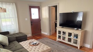 A living room with a sectional sofa, a tufted ottoman, a flat-screen TV on a cabinet, a patterned rug, and a wooden floor. - Charming Stylish & Cozy Coastal Retreat (Rockport)