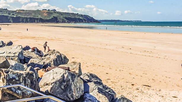 Une plage avec de gros rochers au premier plan, une étendue de sable et un paysage vallonné en arrière-plan.
