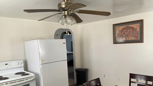 Una cocina compacta con nevera blanca, estufa y ventilador de techo. Una mesa con sillas se encuentra debajo de un cuadro colgado en la pared.