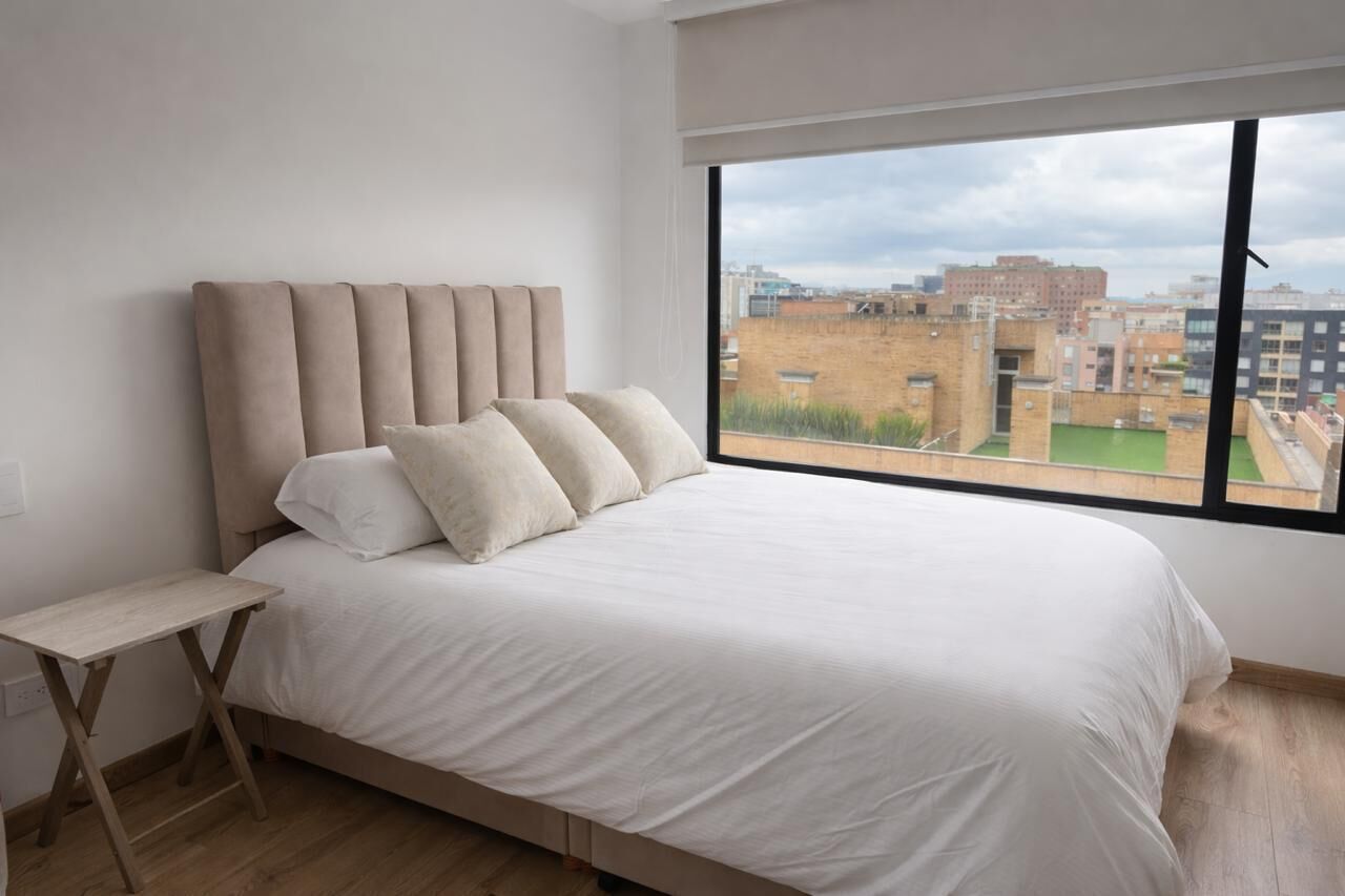 Une chambre moderne avec un grand lit, une petite table de chevet et une vue sur les immeubles par une grande fenêtre.