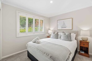 Ein Schlafzimmer mit einem großen Bett, einem Fenster mit Rollläden, einem Nachttisch mit Lampe und einem gerahmten Bild an der Wand.