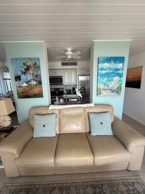 A living room with a beige leather sofa, two light blue cushions, and a patterned rug. There are two paintings on the walls, one depicting palm trees and the other a beach scene. A ceiling fan is visible above the sofa. - Lovely renovated 2br/ 2 ba condo in Siesta Key  across from the #1 Beach in USA (Siesta Key)