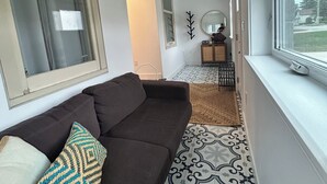 Ein schmaler Flur mit einem dunkelbraunen Sofa, einem Teppich mit Muster und einem Fenster mit Blick auf Bäume.