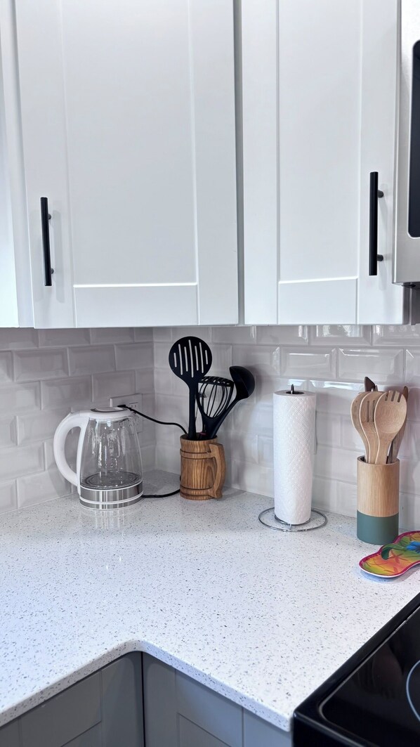 Een keuken met witte kasten, een sproetjesprint aanrecht, een waterkoker, een houten keukengerei houder, een keukenrol en houten lepels.
