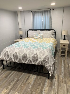 Une chambre à coucher avec un lit, deux tables de chevet, une fenêtre avec des rideaux et un plancher en bois.