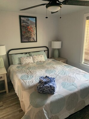 Un dormitorio con cama, ventilador de techo, mesita de noche con lámpara, un cuadro en la pared y una ventana con persianas.