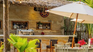 Un espace bar avec des chaises en bois et un parasol blanc, entouré de plantes tropicales et d’un mur en pierre.