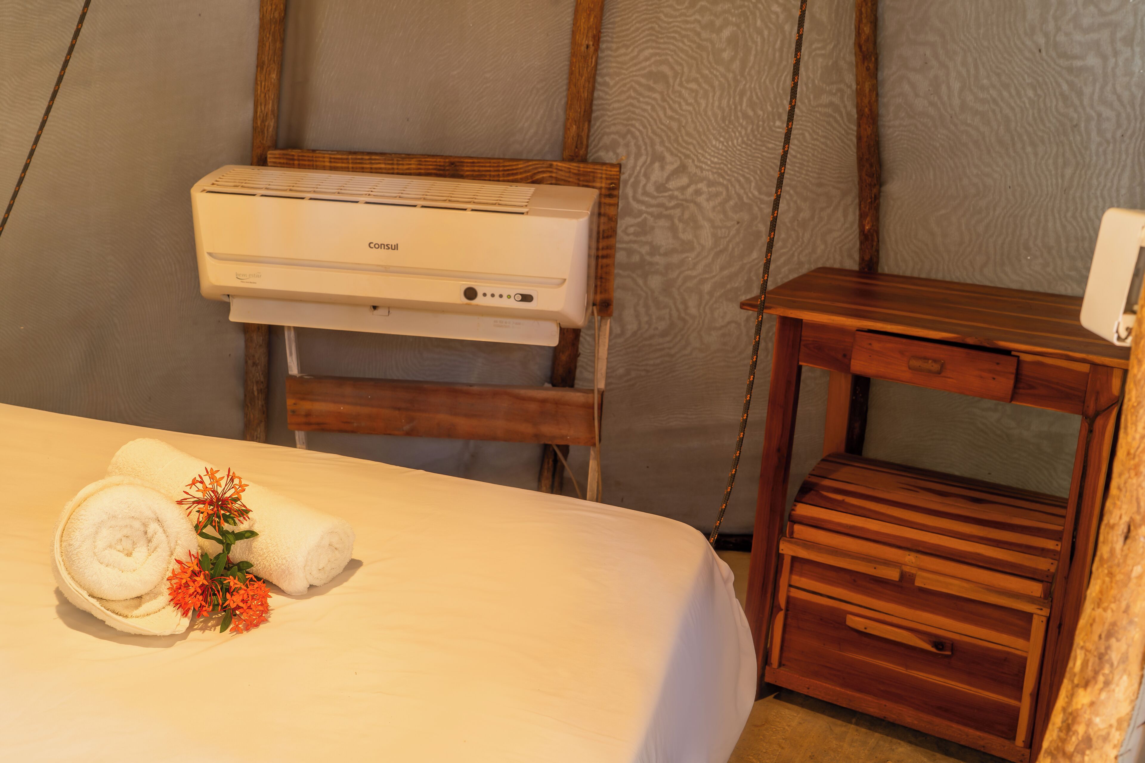 Una habitació d'hotel amb un llit, una petita taula de nit de fusta, aire condicionat fixat a la paret i un calaixera de fusta.