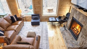 A cozy living room with a stone fireplace, leather sofas, and a view of the outdoors through large windows. - Restful Haven - Hot Tub at Secluded Modern Cabin | Private Trails (Altamont)