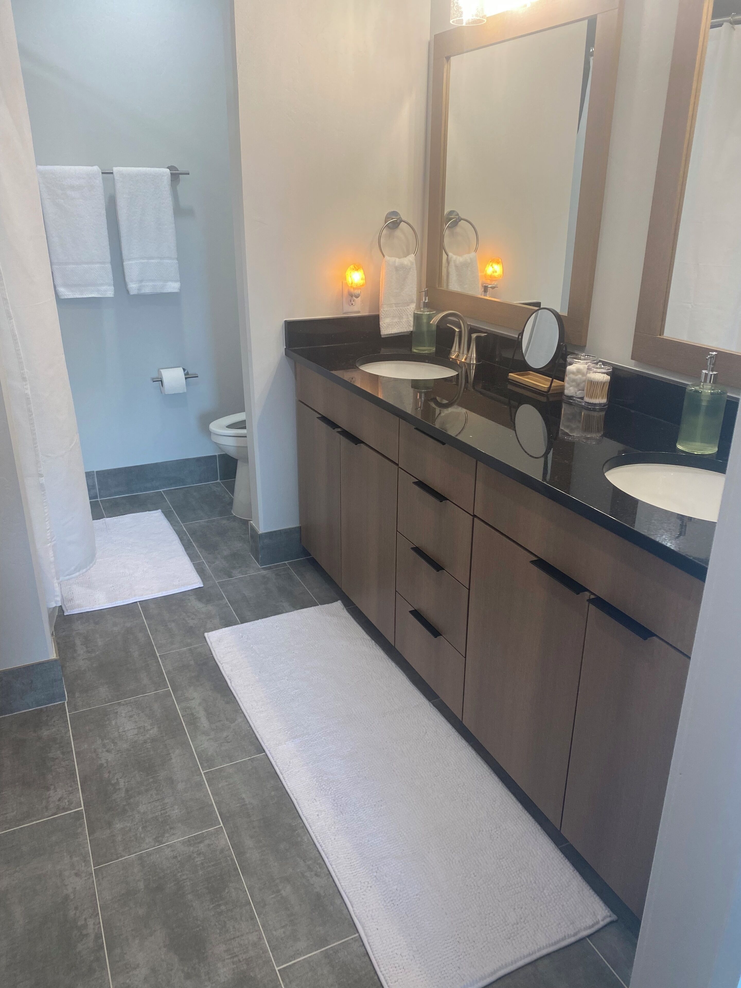 A modern bathroom with a black countertop, two sinks, a large mirror, and a toilet.