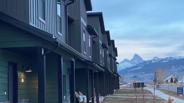 A row of modern buildings with balconies, a stone foundation, and a view of mountains in the background.