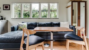 Un salon moderne avec un canapé en cuir noir, une table basse en bois et deux chaises en bois. Une grande fenêtre offre une vue sur la verdure à l’extérieur.