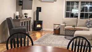 A living room with a wooden dining table, black chairs, a sofa, a fireplace, and a large window. - Spacious new 2 bedroom home in charming Bass Harbor (Tremont)