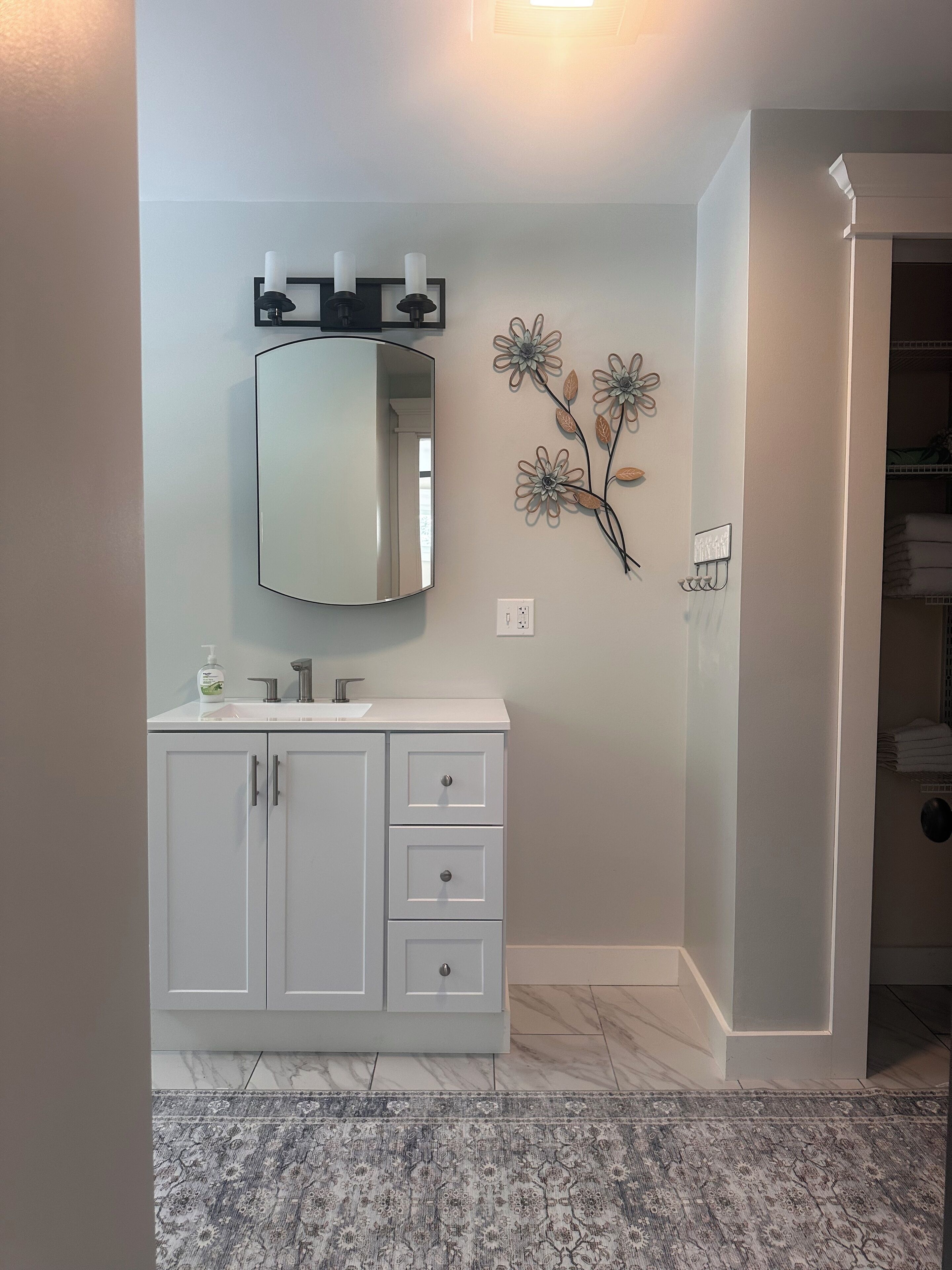 Un baño con tocador blanco, un espejo, una lámpara de pared y un arreglo floral decorativo.