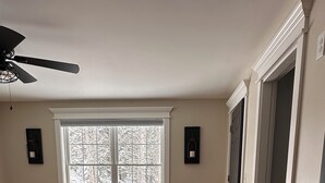 A bedroom with a large window, wooden flooring, a bed with a floral bedspread, a ceiling fan, and two wall-mounted light fixtures. - Spacious new 2 bedroom home in charming Bass Harbor (Tremont)