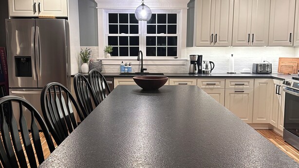 A modern kitchen with a central island, black chairs, white cabinets, and a hanging light fixture. - Spacious new 2 bedroom home in charming Bass Harbor (Tremont)