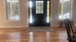 A spacious room with a wooden floor, a black door, and two windows with a view of trees. - Spacious new 2 bedroom home in charming Bass Harbor (Tremont)