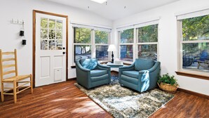 Une pièce avec un plancher en bois, deux fauteuils bleus, une chaise en bois, une porte blanche et de grandes fenêtres donnant sur des arbres et du mobilier d’extérieur.