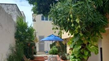 Un patio con una fuente de piedra, un árbol de hojas verdes, una sombrilla azul y un edificio blanco.