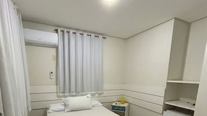 A hotel room with a bed, a bedside table, a nightstand, a small refrigerator, and a window with curtains.