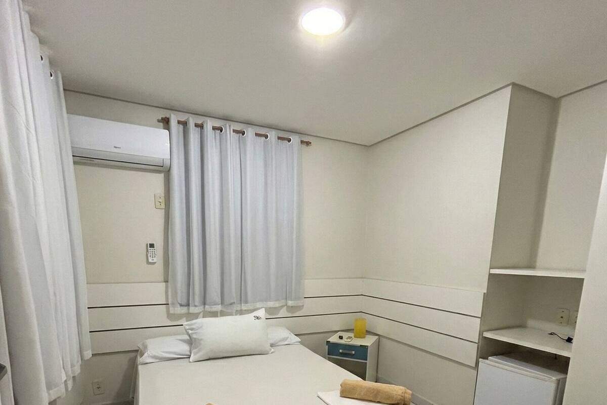 A hotel room with a bed, a bedside table, a nightstand, a small refrigerator, and a window with curtains.