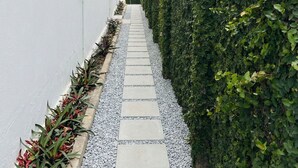Ein schmaler Weg, begrenzt von weißen Mauern und einem Hecken, der zu einem Gebäude mit einem sichtbaren Fenster und Klimaanlage führt.