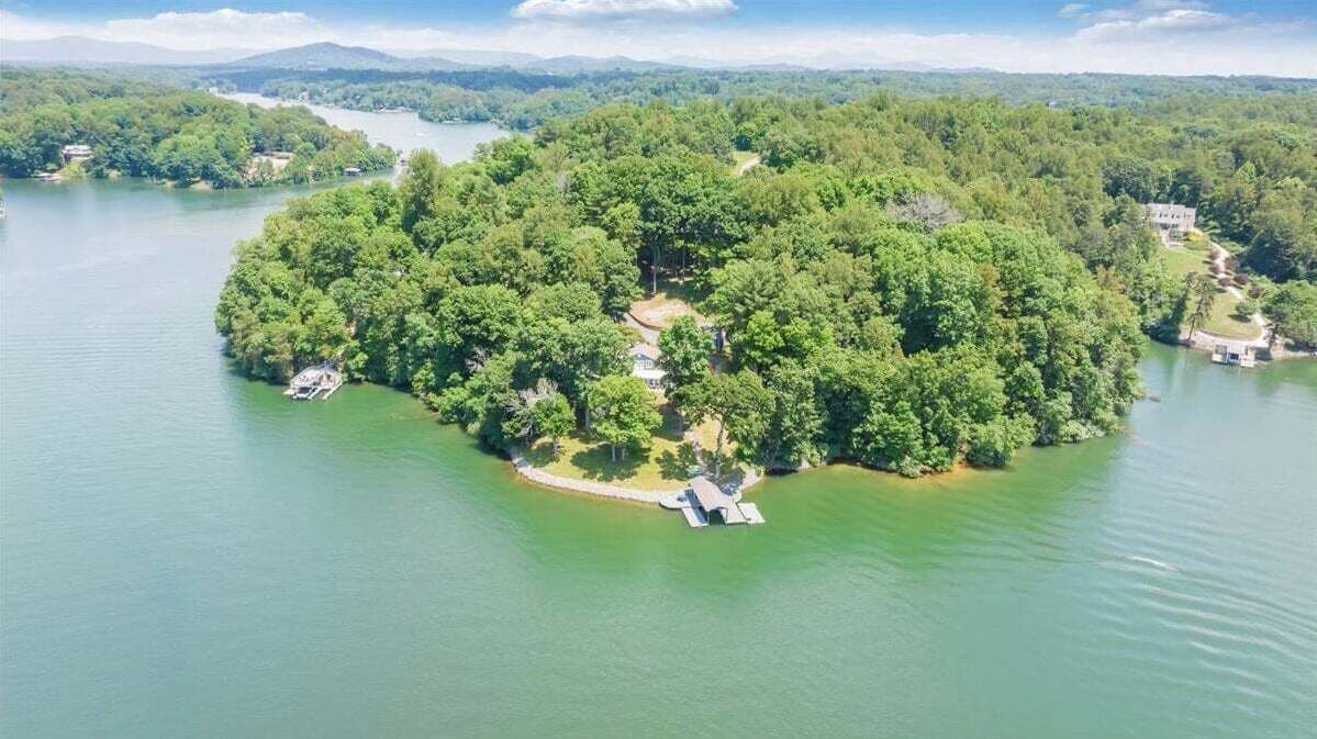Ein Luftbild einer kleinen Insel mit dichtem Grün und einigen Gebäuden, umgeben von ruhigem Wasser.