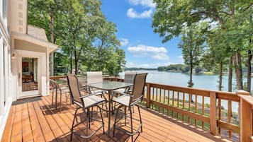 A wooden deck with a glass table and chairs, overlooking a lake with trees and buildings in the distance.
