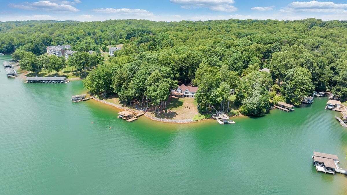 An aerial view of a lakeside property with a house surrounded by dense trees, docked boats, and a pier.