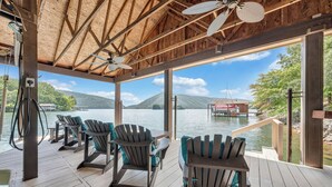A wooden deck with Adirondack chairs overlooking a lake, a thatched roof, and a view of a dock and mountains. - Lake Allure - Private Dock, Indoor Pool, and Hot Tub (Huddleston)
