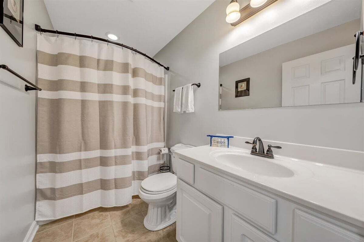 Un baño con cortina de ducha a rayas, un inodoro blanco, un lavabo blanco con un espejo encima y una toalla colgada en la pared.