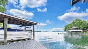 Ein Holzsteg mit einem Boot, einem überdachten Bereich und Blick auf einen See mit Bäumen und einer entfernten Uferlinie.