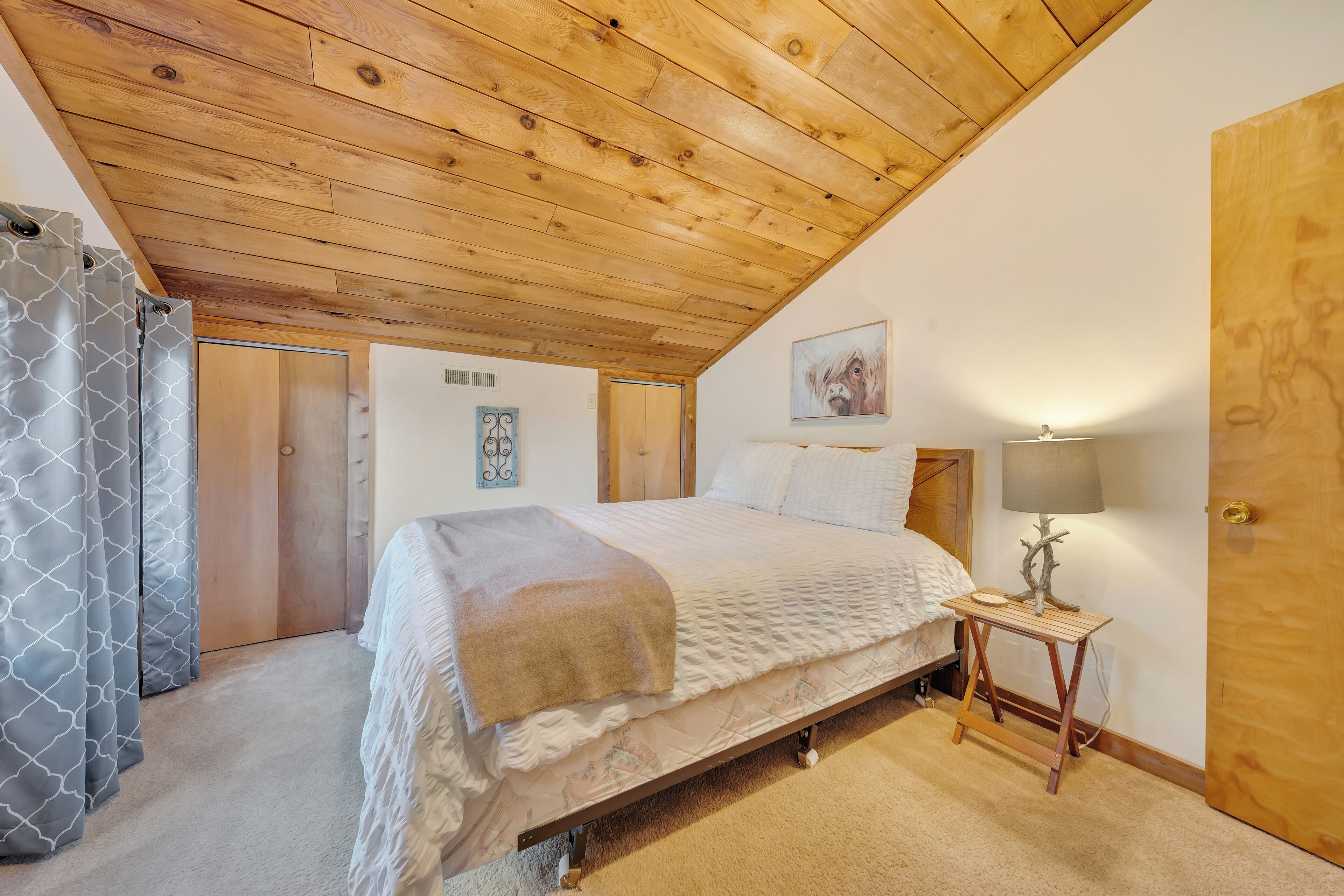 Ein Schlafzimmer mit Holzbalkendecke, einem Bett mit weißer Bettdecke, einem Nachttisch mit Lampe und einer Tür.