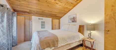 Une chambre avec un plafond en bois, un lit recouvert d’une courtepointe blanche, une table de chevet avec une lampe et une porte.