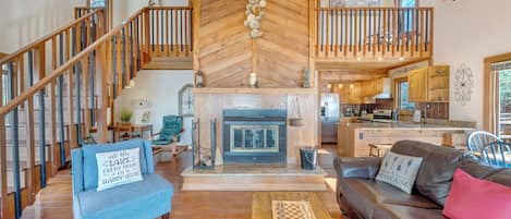A spacious living area with a fireplace, wooden flooring, a staircase, and a kitchen in the background.