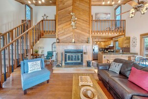 Un amplio salón con chimenea, suelo de madera, un sillón azul, un sofá de cuero marrón y una escalera de madera.