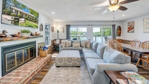 A spacious living room with a fireplace, a large sofa, a dining table, and a ceiling fan. - Lakeside Sunsets - Private Dock, Hot Tub & Firepit (Moneta)
