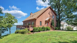 A large, single-story house with a deck overlooking a body of water, surrounded by lush greenery. - Pine Knob Point - Lakehouse w/ Private Dock (Moneta)