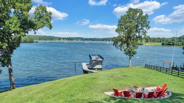 A lakeside deck with a fire pit and red chairs, overlooking a calm lake with a dock and a bridge in the distance. - Pine Knob Point - Lakehouse w/ Private Dock (Moneta)