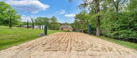 A sandy equestrian arena with a wooden fence, surrounded by green grass and trees.
